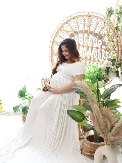 A serene portrait of a mom-to-be, lost in thought while cradling her bump. The white gown and natural props like potted plants and a peacock chair create a peaceful atmosphere.