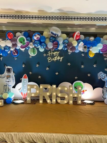 An out-of-this-world space theme for Prish's first birthday. The stage features a starry night backdrop, a large balloon arch with planets, astronaut cutouts, and marquee letters.