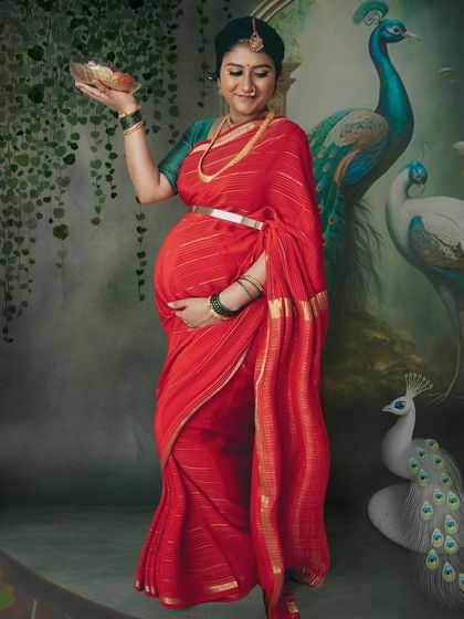 A stunning full-length portrait of the mother-to-be in a traditional red saree, holding a floral offering. The painted peacock background adds a touch of royal elegance to the scene.