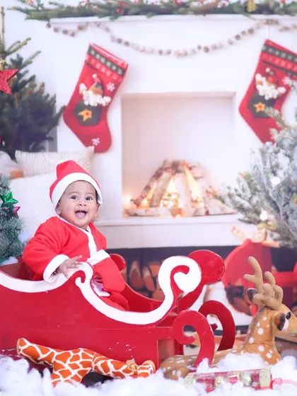 Santa's little helper has arrived. A baby dressed in a Santa suit sits in a red sleigh, surrounded by Christmas trees and snow.