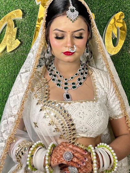 A bride in a white lehenga with a layered emerald and diamond-look necklace. The look is completed with a maang tikka and matching white and green bangles.