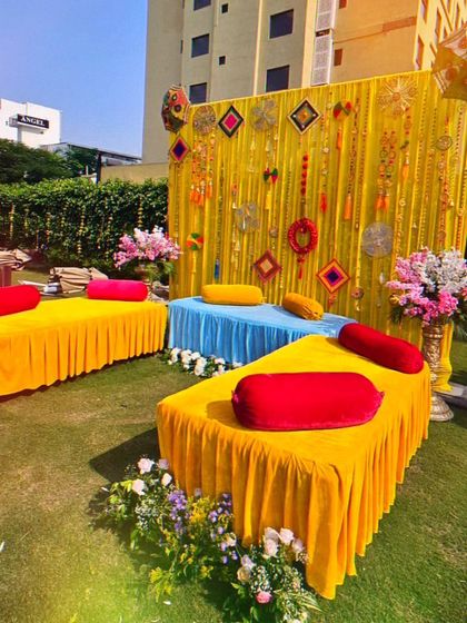 A vibrant outdoor Mehendi seating area with low gaddas in yellow, blue, and red. The backdrop features yellow drapes with traditional hangings and tassels.