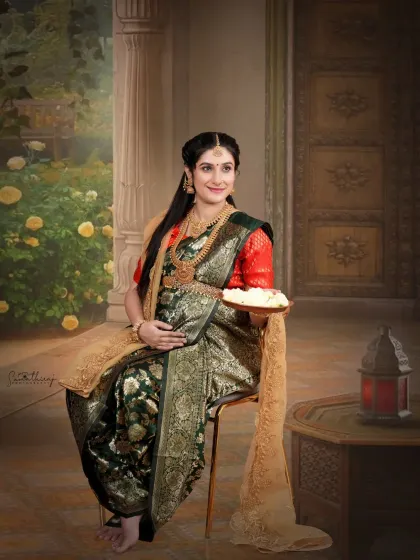 A regal portrait of a mother-to-be in a traditional green saree, holding a plate of flowers. The classic indoor setup with an antique table and lantern adds a touch of royal elegance.