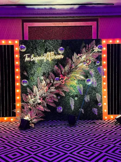 "The Beginning of Forever." This unique photobooth combines a lush green wall with modern bulb-lit frames and a custom neon sign, creating a stylish and meaningful backdrop.