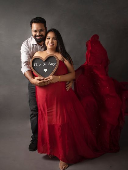 A happy couple announcing "It's a Boy" with a heart-shaped chalkboard. The flowing red fabric of her gown adds a beautiful, dynamic element to the shot.