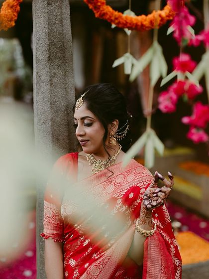 A beautiful portrait of a bride in a red saree, framed by soft foreground elements and colorful flowers.