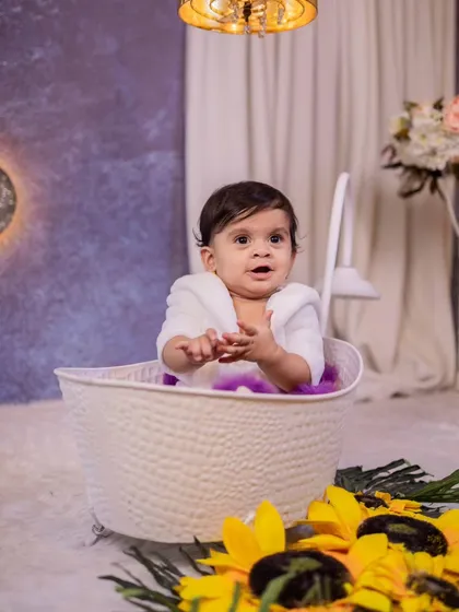 A cute baby wrapped in a towel sits in a basket, part of a fun 'bath time' themed shoot. The sunflowers in the foreground add a bright and cheerful pop of color.