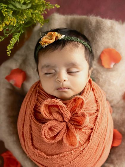 A close-up of a newborn's face, wrapped in a warm orange swaddle. The image focuses on the baby's peaceful expression and the soft texture of the wrap.