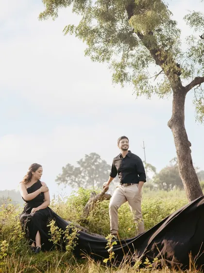 A wide-angle shot from a pre-wedding session, showing the couple in a beautiful, natural landscape. The styling is simple and elegant, with the bride in a flowing black gown, creating a sense of timeless romance.