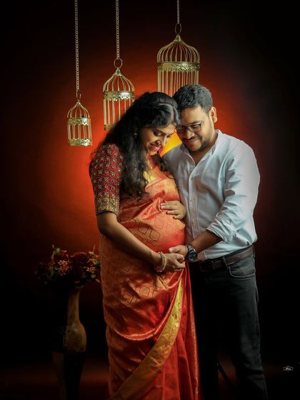 A loving couple admires the baby bump in this warm, intimate studio portrait. The dramatic lighting highlights the rich colors of the traditional red saree.