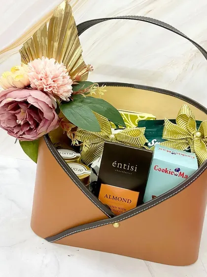 A classic Roka hamper in a tan leatherette envelope basket, filled with gourmet treats and adorned with a beautiful floral arrangement.