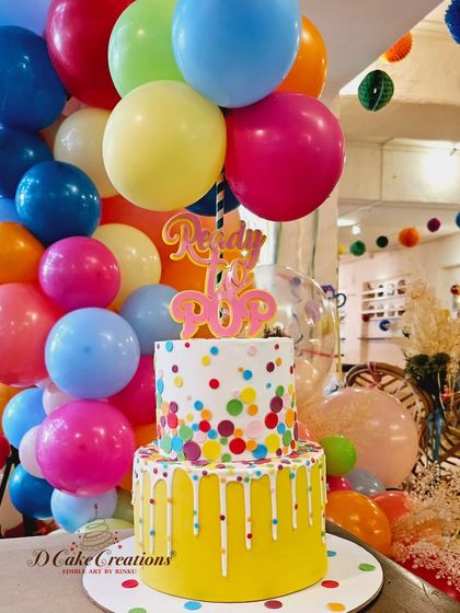 The "Ready to Pop" baby shower cake, a two-tier creation with a bright yellow drip and colorful confetti, topped with real balloons.