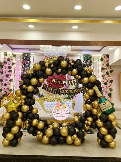 An elegant and grand retirement party decoration in a banquet hall. The setup features a large circular arch with classic black and gold balloons, a 'Happy Retirement' banner, and a champagne bottle foil balloon for a celebratory touch.