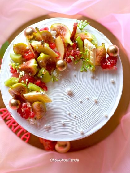 A beautiful fresh fruit cake, topped with a mix of seasonal fruits like figs, kiwi, and pomegranate. The fruit is arranged artfully on a base of white cream with a textured finish.