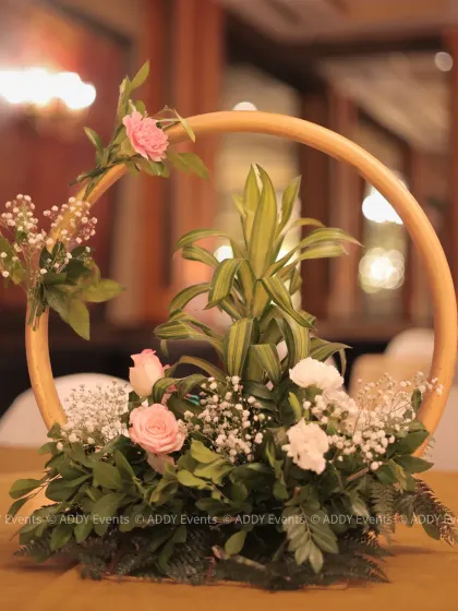 A modern floral centerpiece featuring a golden hoop with an arrangement of pink roses, white carnations, and fresh greenery. This design adds a touch of contemporary elegance to guest tables at a corporate dinner.