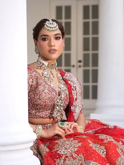 A seated portrait that captures the grandeur of the red lehenga. The styling emphasizes the layered jewelry and the rich, detailed fabric of the outfit.