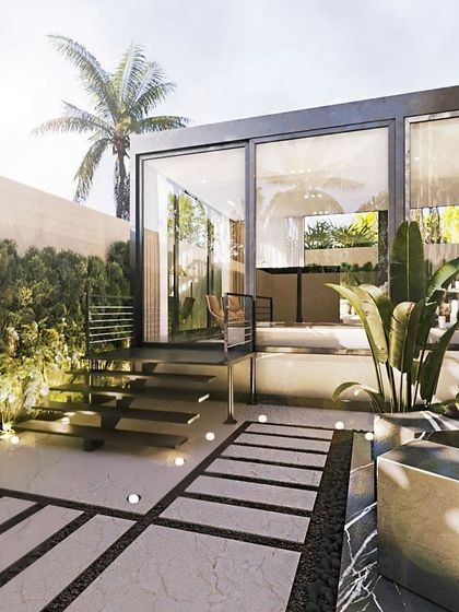 This daytime view shows a glass-encased patio lounge set in a lush garden. The design includes a paved walkway with ground-level lighting, leading up to the elevated structure.