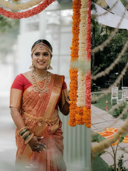 A bride stands smiling during her morning wedding ceremony. The traditional floral decorations and the green lawn create a vibrant and classic South Indian wedding scene.