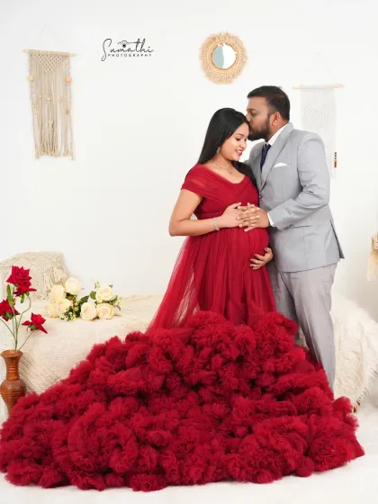 A romantic couple portrait with the husband embracing his wife from behind. Her stunning red gown and the boho-chic studio setup create a warm and stylish scene.