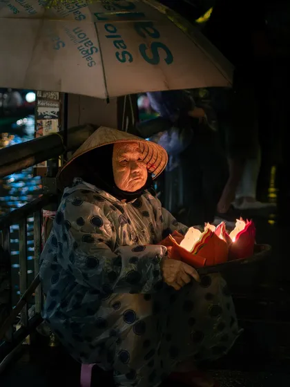 A poignant portrait of an elderly Vietnamese woman in a traditional conical hat, selling candle-lit lanterns from a boat at night.