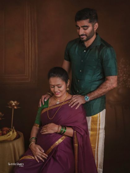 A quiet, traditional moment between a couple. The partner stands behind his seated wife, who is dressed in a purple saree, his hands gently resting on her shoulders.