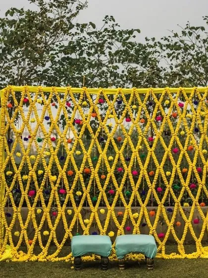 This is a simple yet beautiful photo backdrop for an outdoor haldi event. I created a net of yellow flower strings, dotted with colorful pom poms, which looks lovely against the natural green background.