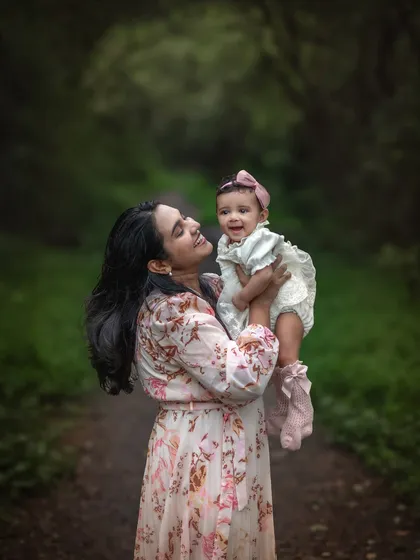 A mother joyfully lifts her smiling baby into the air, her face filled with love and happiness. Set against a lush green background, this image freezes a perfect, fleeting moment of connection.
