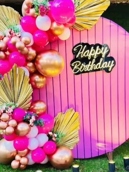 A close-up of a vibrant and trendy birthday backdrop. I used a circular pink panel with a custom "Happy Birthday" neon sign, accented with hot pink and chrome gold balloons and dried palm leaves for a modern look.