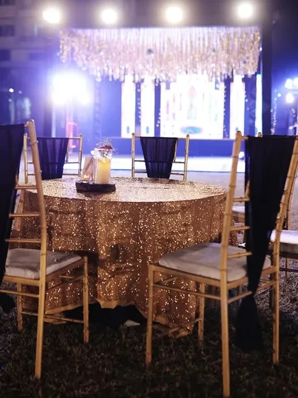 A guest table at a Sangeet, with a gold sequin tablecloth and elegant black chair drapes, showing the stage in the background.