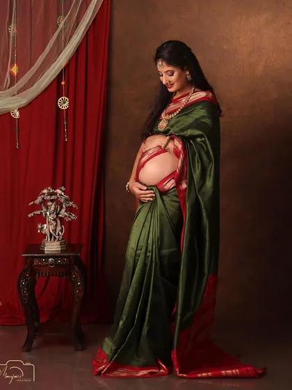 A beautiful solo portrait in a green and red saree, her hand lovingly on her bump. The traditional setup is rich and elegant.