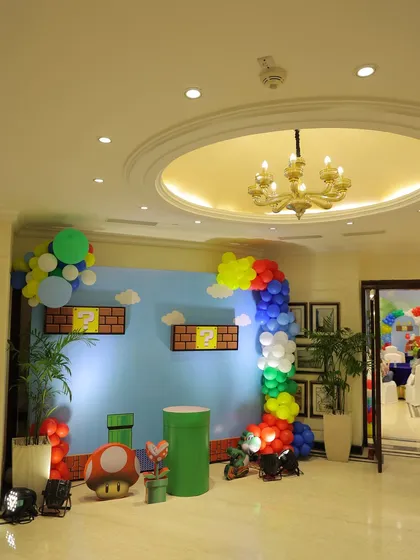 A full view of the Super Mario themed stage. The setup includes a large backdrop of the game's sky, warp pipes, a mushroom prop, and a rainbow balloon arch, creating an immersive gaming world.