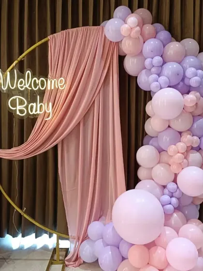 An elegant and minimalist welcome baby setup. A golden ring stand is draped with pink fabric and adorned with a pastel balloon garland and a "Welcome Baby" neon sign.