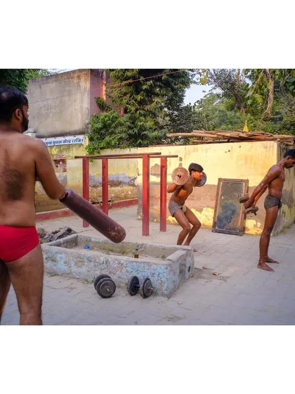 Men training at a traditional 'akhada' or outdoor gym. The scene shows various forms of exercise, from lifting weights to using traditional wooden clubs (mudgar).