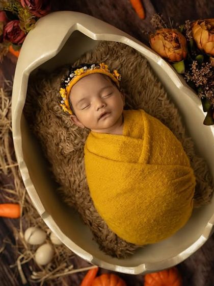 A creative newborn shot where the baby is swaddled in yellow and sleeping inside a large eggshell prop, surrounded by carrots and hay.
