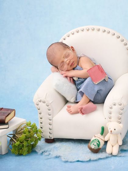 A tiny scholar, this newborn wears little glasses and sleeps in a miniature armchair with a book, a tiny coffee cup, and a teddy bear.