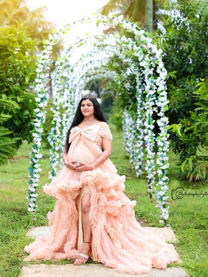 A princess-like portrait of a mother-to-be in a peach ruffled gown and crown, standing under a floral archway.