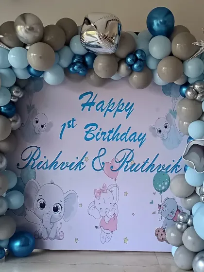 A cute elephant theme for a twins' first birthday. The backdrop features adorable cartoon elephants, framed by a balloon arch in soft blue and grey tones with silver accents.