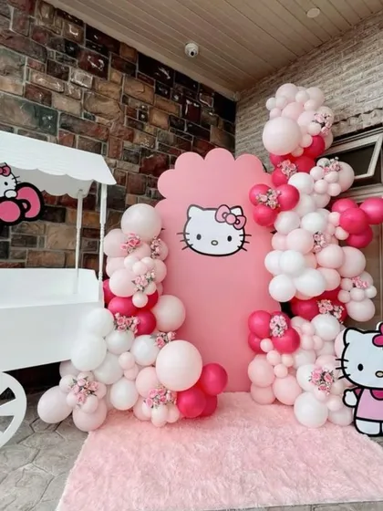 A cute and charming Hello Kitty themed photo corner. This setup includes a pink backdrop, a white candy cart prop, and a beautiful balloon garland in shades of pink and white.