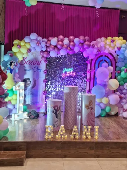 A magical Disney Princess theme for Baani's first birthday. The stage is decorated with a sequin backdrop, character cutouts of Ariel and other princesses, and pastel-colored balloons.