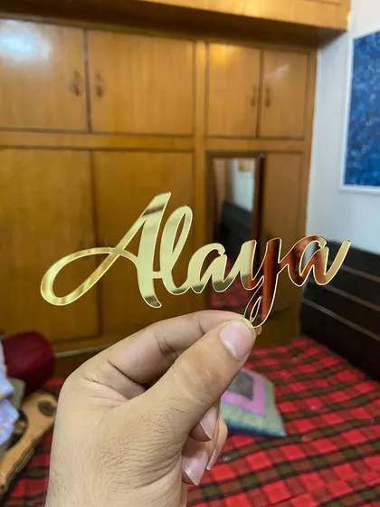 A simple "Alaya" name topper in classic gold, held in hand to show its delicate script and reflective shine.