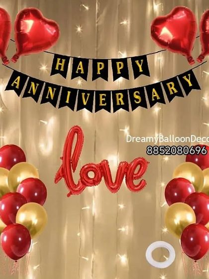 A classic anniversary decoration kit. This setup includes a 'Happy Anniversary' banner, 'Love' foil balloon, and bunches of red and gold balloons, all set against a curtain of warm fairy lights.