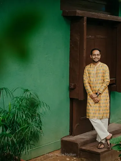 The groom, in a yellow kurta, stands by a doorway, the green foliage and natural light creating a fresh and vibrant portrait.