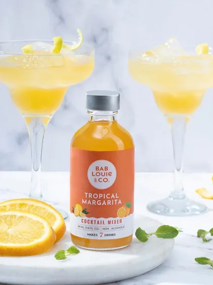 A bright and refreshing shot of Bab Louie & Co.'s Tropical Margarita mixer. The image, complete with prepared margaritas and fresh orange slices, is perfect for a summer beverage campaign.