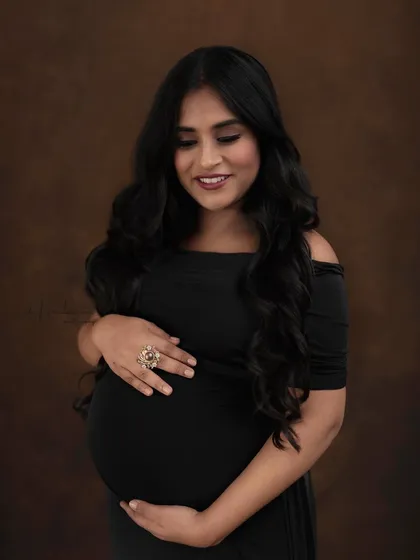 A close-up portrait against a warm, brown backdrop. The focus is on her beautiful, glowing smile and the simple elegance of the off-shoulder black dress.