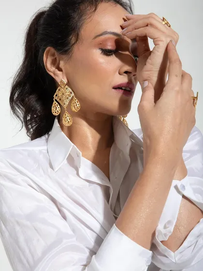 A close-up from the studio shoot, focusing on the delicate gold earrings and the model's expressive hands. The clean lighting and simple white shirt ensure the accessories and makeup are the stars.