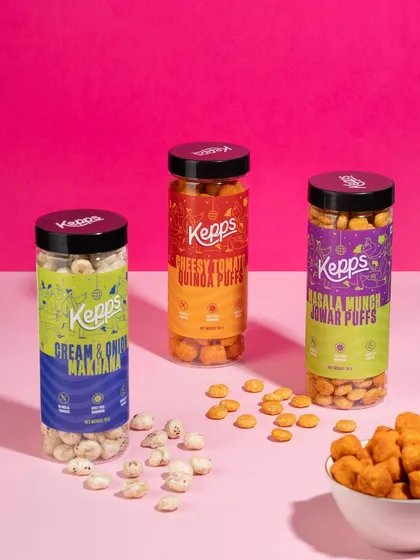 This image showcases the full range of snack flavors against a vibrant pink background. The clean arrangement allows the colorful packaging to stand out, while the scattered puffs add a touch of texture and playfulness.