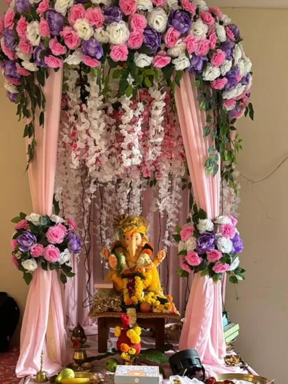 A lovely pastel-themed Ganpati mandap with pink, purple, and white flowers creating a canopy. The hanging wisteria and soft pink drapes add to the dreamy and gentle aesthetic.
