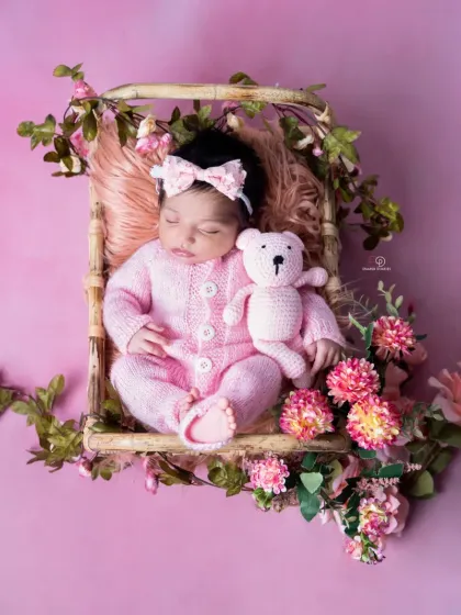 A newborn girl in a pink knitted romper sleeps in a bamboo basket decorated with pink flowers and greenery. This portrait combines rustic elements with soft, feminine colors.