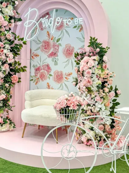 A charming "Bride to be" photo corner with a pink arched backdrop and a floral-print interior. The setup is completed with a plush white chair, a decorative white bicycle, and an abundance of pink roses.