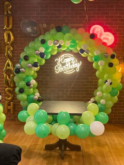 A football-themed birthday backdrop in a restaurant. The circular balloon arch in shades of green is accented with footballs and a neon sign.